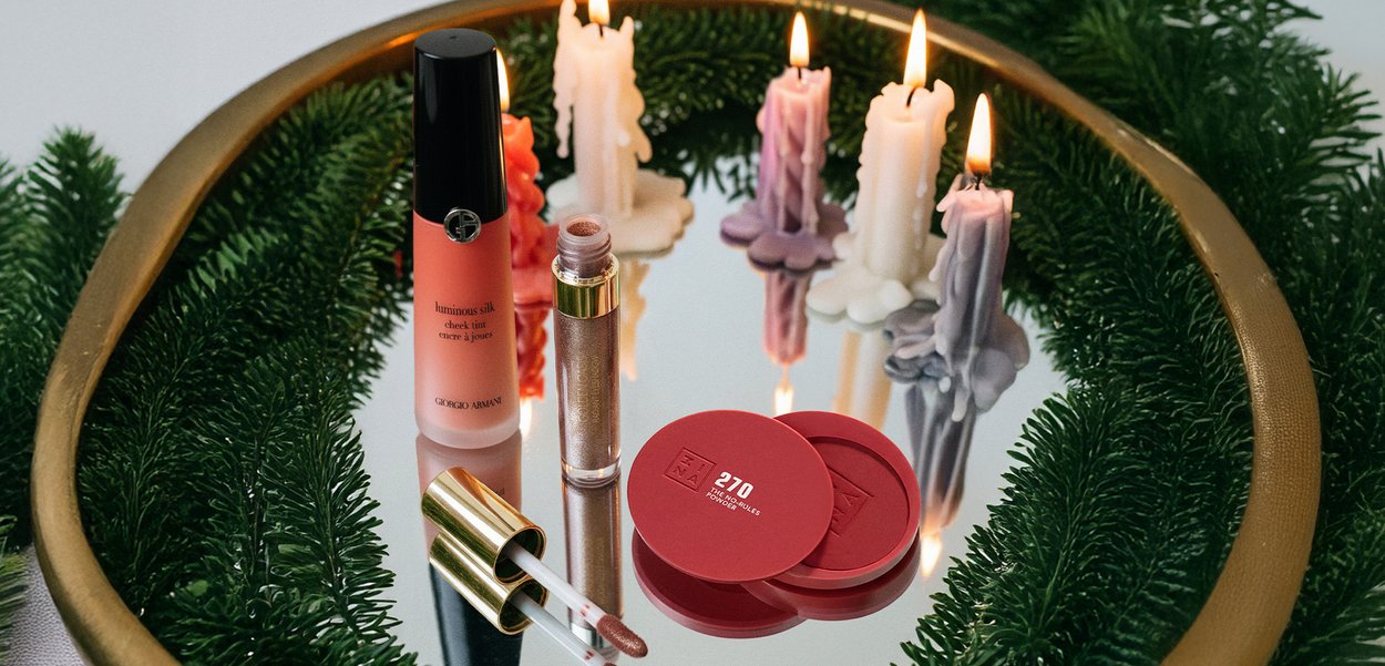 Pyöreä kultainen tarjotin, jossa vihreitä männynoksia, pitelee palavia värikkäitä kynttilöitä sekä sekalaisia punaisia ja kultaisia meikkituotteita applikaattoreineen.