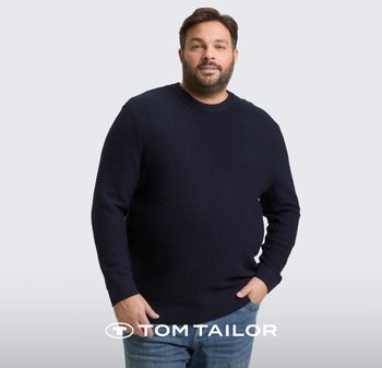 Man die een gestructureerde marineblauwe trui en blauwe spijkerbroek draagt, staand met één hand in zijn zak tegen een lichtgrijze achtergrond.