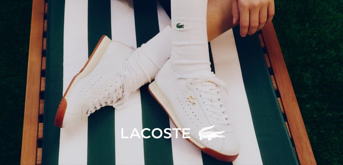 Mujer acostada sobre el césped con shorts negros y camiseta blanca, con zapatillas blancas de Lacoste colocadas cerca sobre el césped.