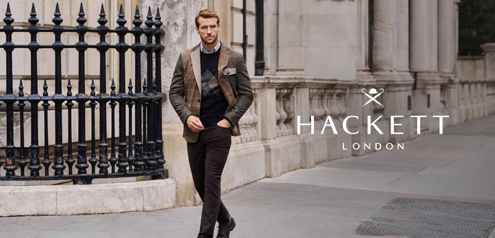 Mann, der auf einem Stadtweg geht, trägt einen Patchwork-Blazer, einen gemusterten Pullover, dunkle Hosen und schwarze Schuhe, in der Nähe eines Eisenzauns und eines Steingebäudes.