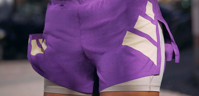 Personne portant un short de sport violet avec des motifs de bandes beiges sur un short de compression blanc, debout à l'extérieur dans une rue urbaine floue.