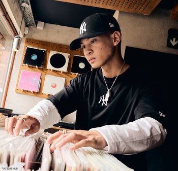 Cappello nero dei New York Yankees, t-shirt nera con logo, maglietta bianca a maniche lunghe sotto, catena d'argento. Mani che sfogliano dischi in vinile.