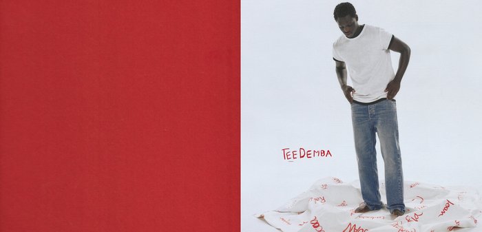 White cotton t-shirt with black accents, paired with light blue denim jeans. The model stands on a white fabric with red text.