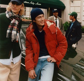 Rote Puffjacke, marineblauer Pullover, hellblaue Jeans. Model mit lockigem blonden Haar trägt einen karierten Schal. Gruppe im Freien neben einem schwarzen Auto.
