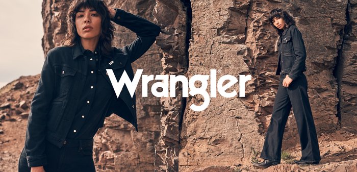 Svart denimjakke over en svart skjorte med knapper, sammen med svarte vide bukser og støvler, satt mot en steinete bakgrunn. Wrangler-merke er synlig.