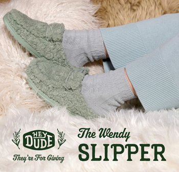 Pantofole morbide verdi testurizzate indossate con calzini grigi chiari e pantaloni blu chiaro, poggiate su un soffice tappeto crema.