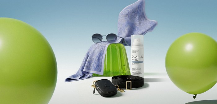 Vihreä hyytelövuoka aurinkolaseissa, violetti pyyhe, hiustenhoitosuihke, musta vyö ja pussukka, kahden vihreän ilmapallon ympäröimänä valkoisella pinnalla.