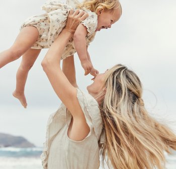 A child in a floral patterned cream romper is held aloft by an adult wearing a light-colored sleeveless top, with ocean waves in the background.