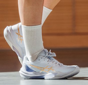 Persoon draagt witte sportschoenen met gouden accenten en witte geribbelde sokken, staand op een gymvloer met een houten muur op de achtergrond.
