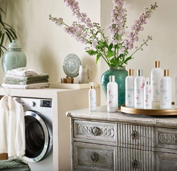 Klare glasflasker med blomsterdesigns, guld- og sølvkapsler på et træbord. Lilla blomster i en grøn vase, vaskemaskine i baggrunden.