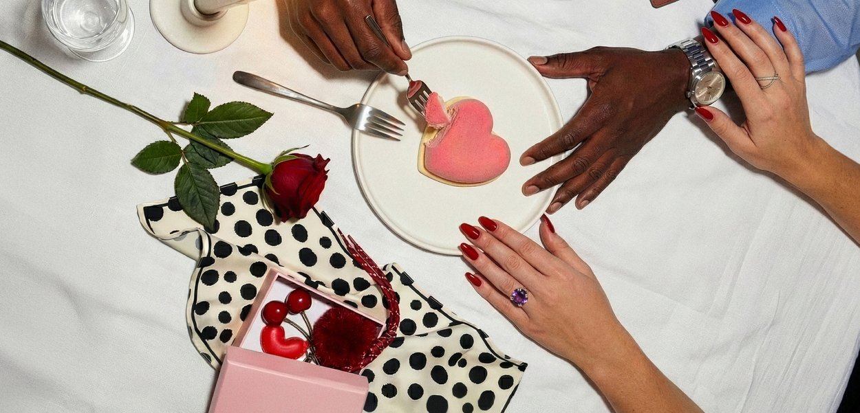 Kaks kätt, üks tumedama ja teine heledama nahaga, plaadi lähedal, millel südamekujulise magustoidu, punase roosi, kinkekarbiga ja musta täpilise kangaga valgel laual.