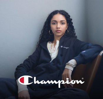 Giovane donna con capelli ricci, che indossa una felpa nera Champion e una maglietta color crema, seduta contro pareti grigie, con sovrapposizione del logo Champion.