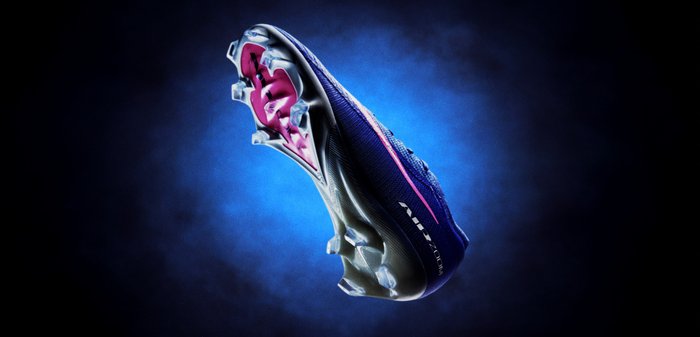 Ποδοσφαιρικό παπούτσι με μπλε σκούρα επάνω επιφάνεια με υφή, ροζ λεπτομέρειες, και διάφανη σόλα με πολλές εξογκώματα για πρόσφυση.