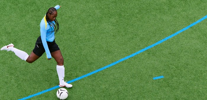 Ženska nogometašica v modrem dresu in belih nogavicah brca črno-belo žogo na zelenem igrišču z modrimi črtami.