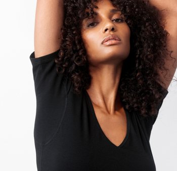 Woman with curly hair raises arms, wearing a black V-neck shirt, looking confidently at the camera against a plain white background.