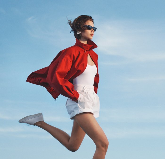Woman in white shorts, tank top, and red jacket with hands in pockets, wearing sunglasses, stepping forward against clear sky.