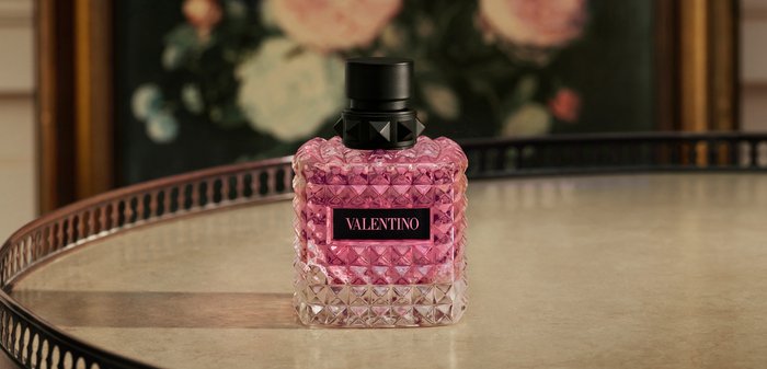 Quadratische pinke Glas-Parfümflasche mit strukturiertem Rautenmuster und schwarzem, nietenbesetztem Verschluss auf einer reflektierenden Oberfläche.