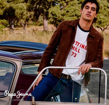 Joven con chaqueta marrón y camiseta blanca con gráfico apoyado en la puerta abierta de un coche convertible vintage al aire libre.