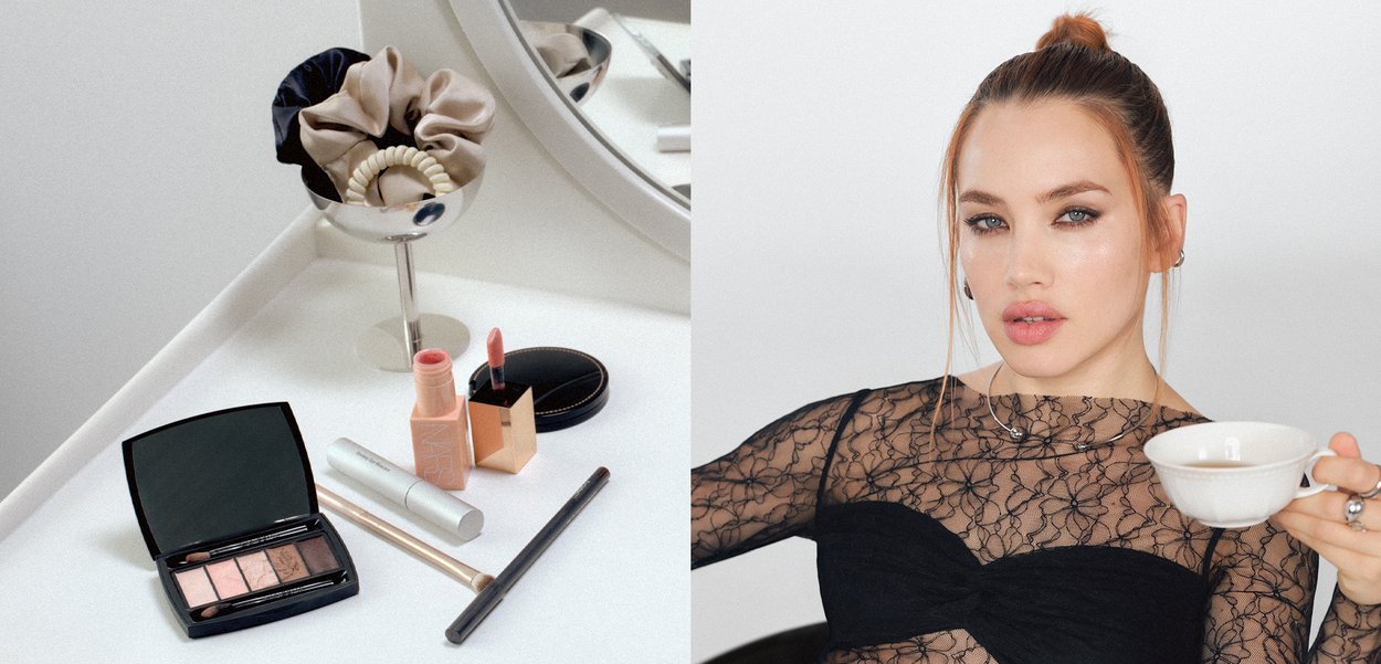 Produits de maquillage sur une surface blanche : palette de fards à paupières noirs, produits pour les lèvres, eye-liner argenté, chouchou en crème dans un porte-métal, et une tasse à thé blanche.