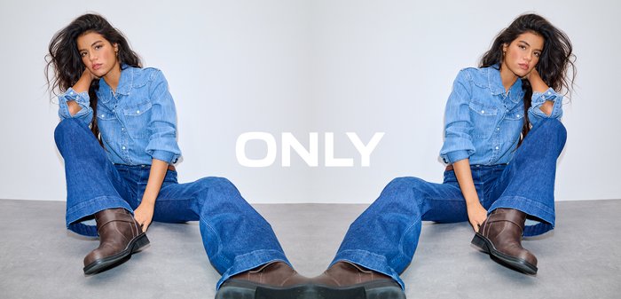 Tânără cu păr lung și întunecat, purtând o cămașă din denim, blugi largi și cizme maro, stând pe un podea gri, sprijinită de un perete alb.