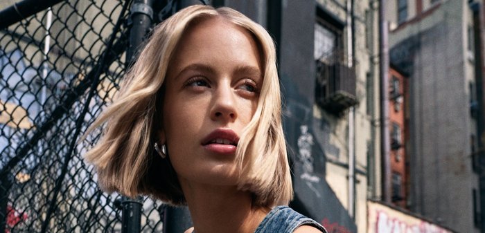 Jeanskleid ohne Ärmel mit tiefem V-Ausschnitt und Knopfdetails. Modell mit schulterlangen blonden Haaren posiert vor einem Graffiti-Hintergrund.