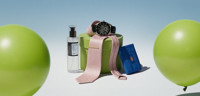 Montre noire sur ruban rose posé sur une boîte ronde verte, flacon transparent de soin de la peau à gauche, portefeuille bleu à droite, deux ballons verts en arrière-plan.