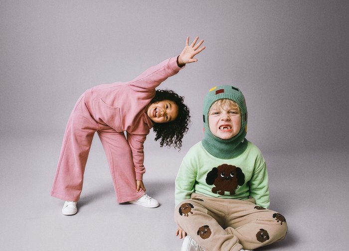 Δύο παιδιά σε γκρι φόντο· το ένα σε ροζ φόρμα τεντώνεται με το χέρι σηκωμένο, το άλλο κάθεται φορώντας πράσινο πουλόβερ με θέμα σκύλου και καπέλο.