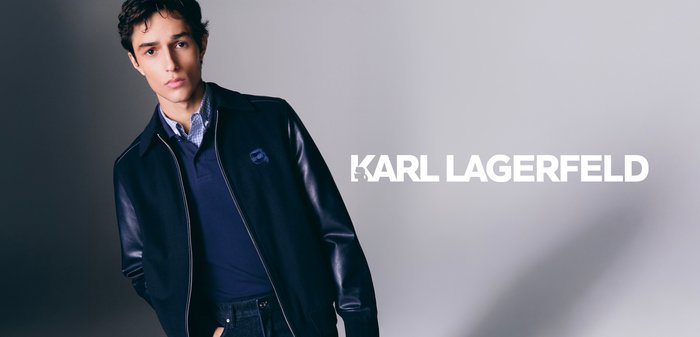 Young man wearing dark jacket with leather sleeves, navy shirt, and jeans, standing against a neutral background with "KARL LAGERFELD" text overlay.