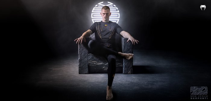 Athletische Figur in einem schwarzen Hochleistungstraininganzug mit goldenen Akzenten, sitzend auf einem strukturierten, dunklen Steinstuhl, minimalistische Hintergrundbeleuchtung.