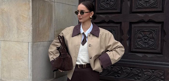 Mujer con chaqueta beige con cuello y puños oscuros, camisa blanca, corbata, pantalones oscuros, gafas de sol, pendientes dorados, sosteniendo un bolso de ante marrón junto a una puerta de madera tallada.