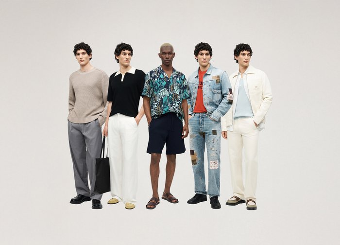 Five men stand side by side wearing diverse casual outfits including knitwear, polo, printed shirt with shorts, patchwork denim, and light jacket with trousers.