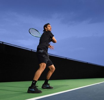 Tennis spiller i svart treningsklær, svinger en racket. Banen har grønn overflate, svart gjerde, under en blå og skyete himmel.