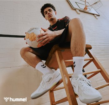 Witte sportschoenen met textuurpatronen, zwart-witte sokken met chevrondesign, en een bruine houten kruk waarop een voetbal ligt.
