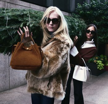 Brun mockaväska och vit hinkväska hålls av två kvinnor. Den ena bär en pälsrock och solglasögon; den andra en tröja med kontrasterande färger.