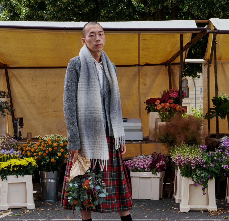 A person wearing a grey cardigan and a red check skirt holds a bouquet, surrounded by vibrant flowers in pots and crates at a market stall.