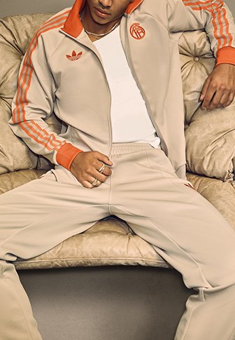 Homme portant un survêtement Adidas beige avec des rayures orange, assis de manière détendue sur un canapé en cuir beige capitonné.