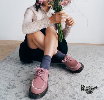 Persona seduta a gambe incrociate su un tappeto, che tiene in mano un bouquet di fiori, indossando scarpe rosa Dr. Martens e calzini grigi.