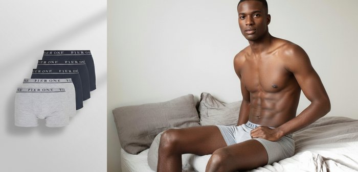 Mladi mišičast moški v sivih boksericah sedi na neurejeni postelji s svetlo sivim posteljnim perilom.