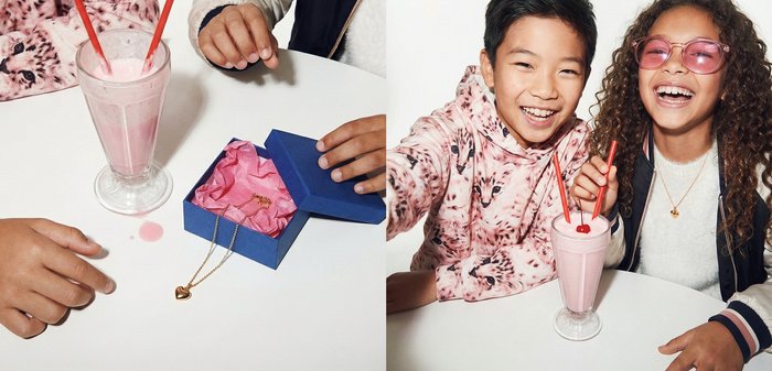 Deux enfants souriant avec un milkshake rose et une boîte cadeau bleue contenant un collier en forme de cœur en or sur une table blanche.