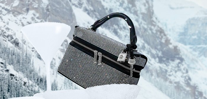Bolso rectangular negro y plateado adornado con pedrería, con dos cremalleras y una asa superior, colocado frente a un paisaje de montaña nevada.