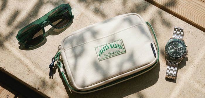 Πράσινα γυαλιά ηλίου με χρώμα, λευκή θήκη Calvin Klein και ασημένιο μεταλλικό ρολόι χειρός με μαύρο καντράν σε ανοιχτή σκυρόδετη επιφάνεια με σκιασμένα μοτίβα.