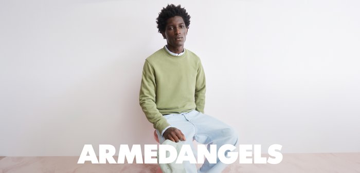 Junger Mann mit dunklem lockigem Haar, der einen grünen Pullover und hellblaue Jeans trägt und vor einem schlichten weißen Hintergrund mit "ARMEDANGELS" Text darunter sitzt.