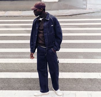 Man klädd i mörk denimjacka och byxor, brun tröja och keps står på ett övergångsställe och tittar åt vänster.