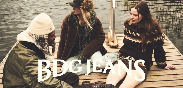 Tre persone sono sedute su una banchina di legno. Uno indossa una giacca blu navy e una sciarpa verde, un altro un cappotto marrone con dettagli in pelliccia, e l'ultimo un maglione a fantasia.