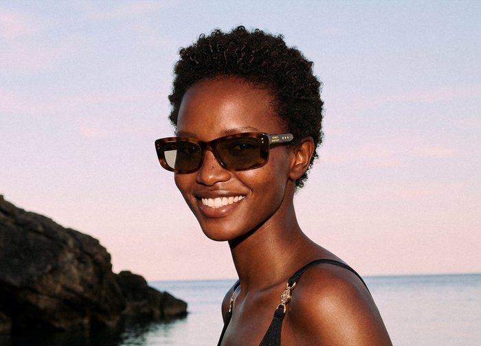 Γυναίκα που χαμογελά με κοντά σγουρά μαλλιά, φοράει γυαλιά ηλίου με σχέδιο χελωνόφρακου και μαύρο τοπ με τιράντες κοντά σε βραχώδη ακτή κατά τη δύση του ηλίου.
