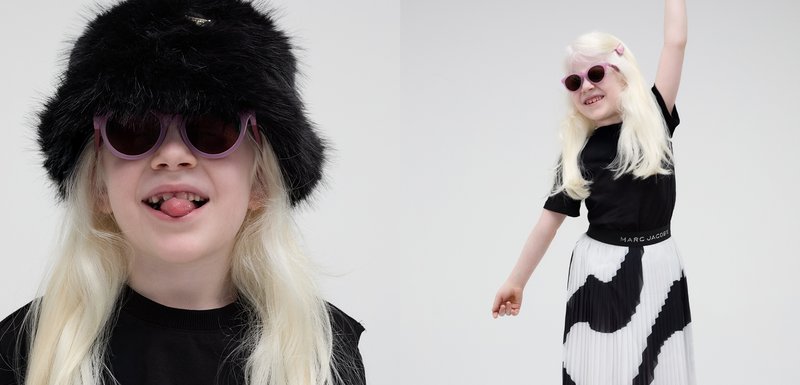 Chapeau seau en fausse fourrure noire, lunettes de soleil rondes violettes, t-shirt noir et jupe plissée blanche avec des motifs abstraits noirs.