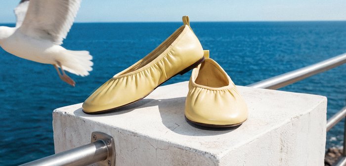Paire de ballerines jaunes reposant sur un rebord en pierre près d'une rambarde métallique avec la mer bleue et une mouette en vol en arrière-plan.