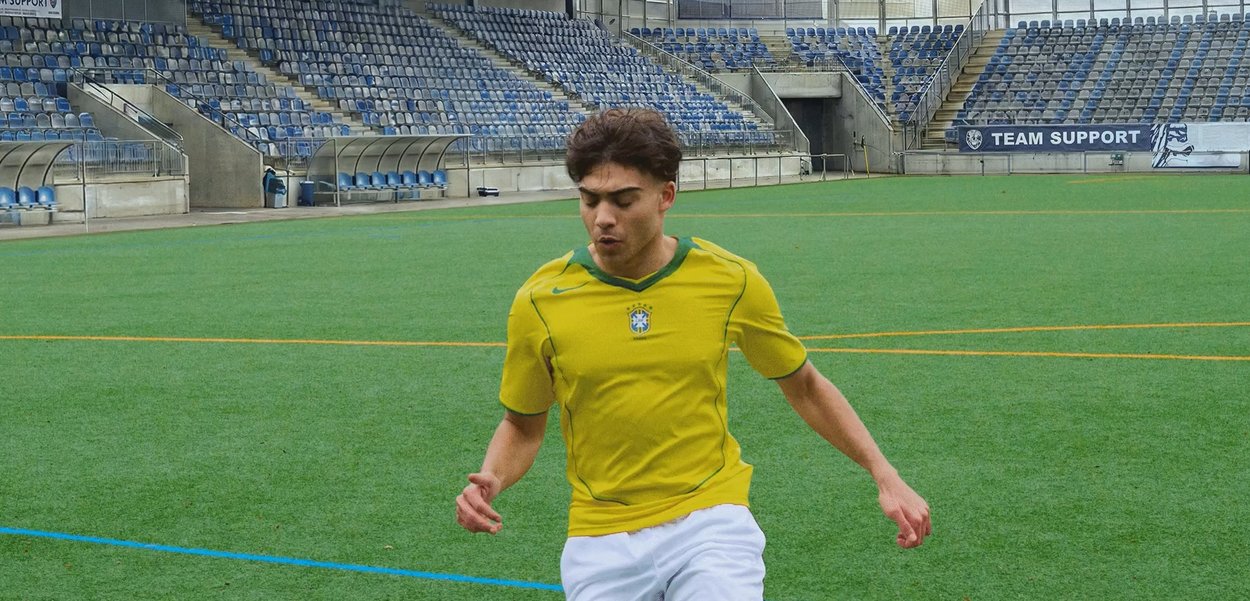 Joven jugador de fútbol masculino con camiseta amarilla y pantalones cortos blancos controlando el balón en un campo verde con asientos vacíos en el estadio.