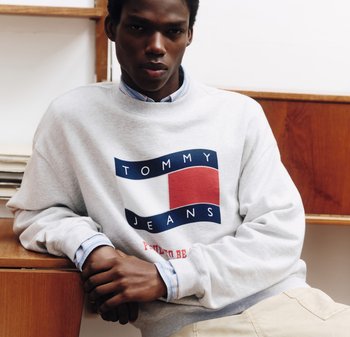 Jonge man met een wit Tommy Jeans sweatshirt en beige broek, leunend op een houten bureau in een binnenomgeving.