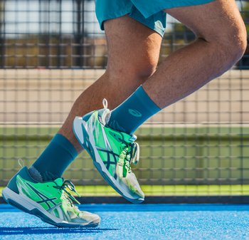 Hardloper in blauwe korte broek, turquoise sokken en groene sportschoenen op een blauw sportveld vlakbij een net.